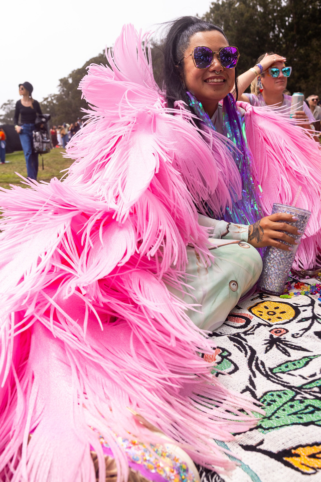 A person wearing a vibrant pink feathered outfit sits on a colorful patterned blanket, smiling and holding a silver tumbler, surrounded by others outdoors.